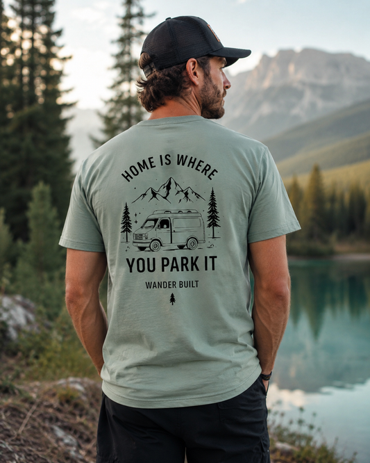 A man wearing a sage green t-shirt with a black graphic print on the back, standing outdoors in front of a mountain lake.