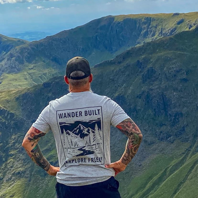 A man stands with his back to the camera, looking out over a vast green mountain valley. He is wearing a light gray t-shirt with a large black graphic on the back that reads 'WANDER BUILT' and 'EXPLORE FREELY', along with a dark baseball cap.